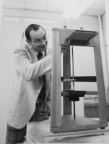 Jennings in a tie and light- colored sports jacket leans over and adjusts a spring suspended from the seismometer frame, a 3-sided structure with a base that sits on a table.
